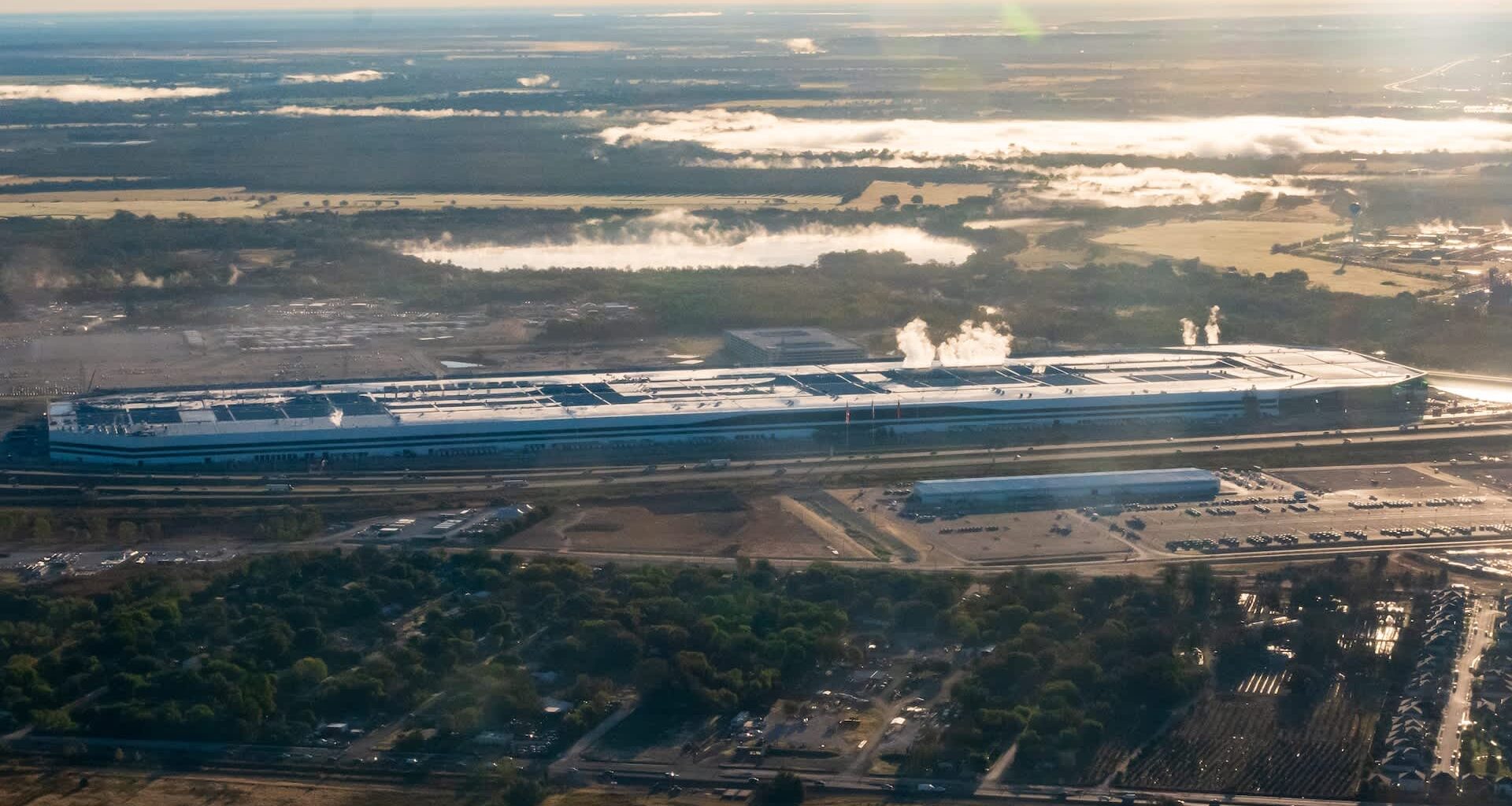 Aerial view of a the Tesla Gigafactory in Austin, Texas, with smoke rising, surrounded by greenery and parking lots.