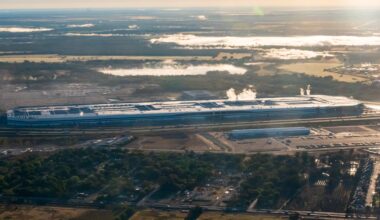 Aerial view of a the Tesla Gigafactory in Austin, Texas, with smoke rising, surrounded by greenery and parking lots.
