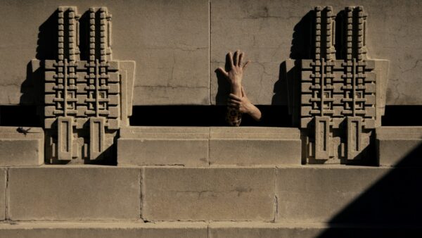 A still image from a video featuring two hands emerging from the base of an architectural structure.