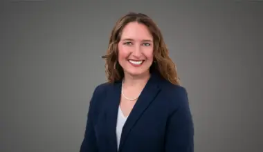 Professional headshot of a smiling woman with curly brown hair in a navy blue suit against a simple gray background