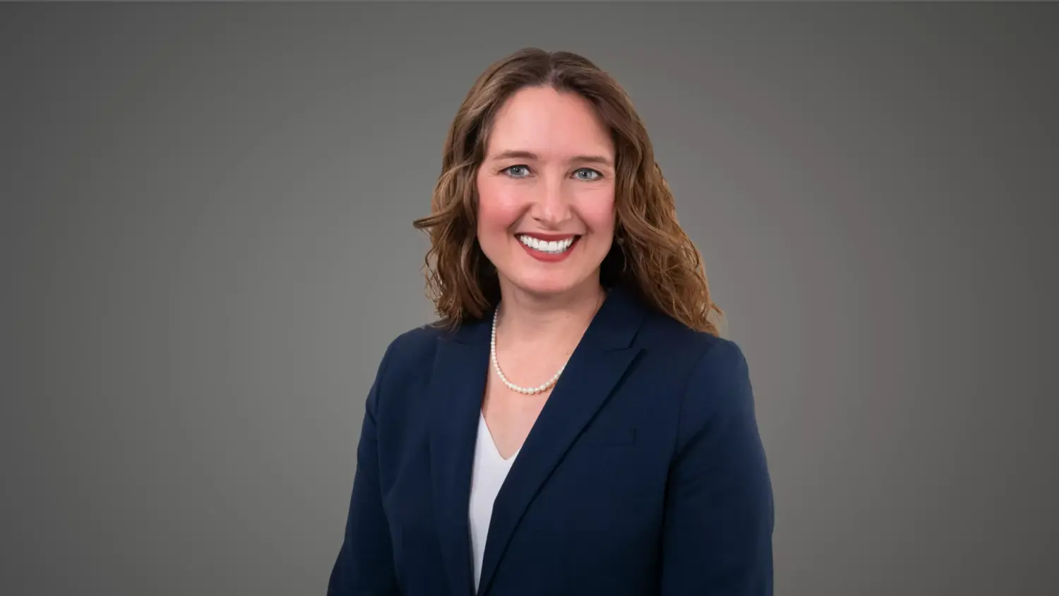 Professional headshot of a smiling woman with curly brown hair in a navy blue suit against a simple gray background