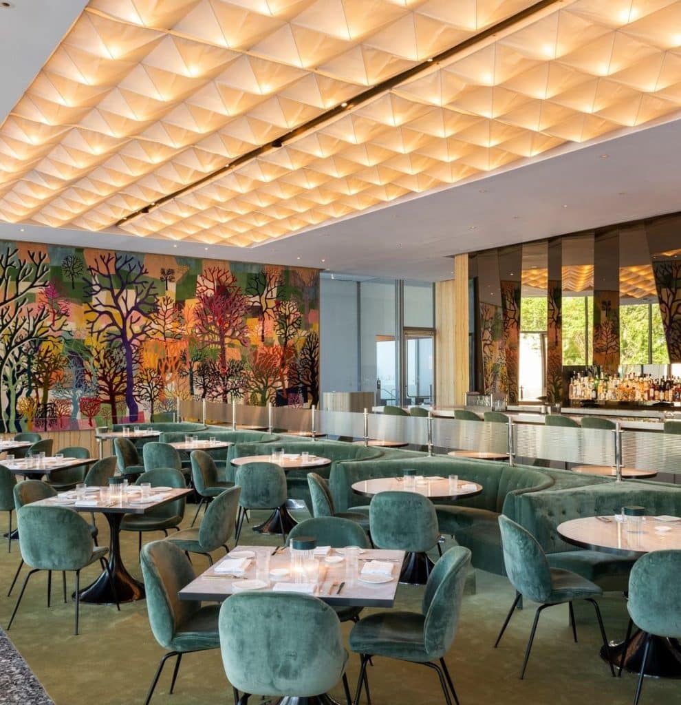 Image shows the interior of Le Jardinier at MFAH with plush velvet chairs, a suspended light installation, and mural in the background.