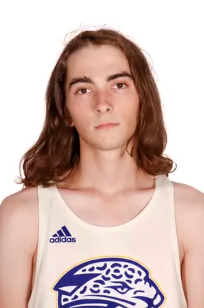 Graycen Vargo, a young man with long brown hair, wearing a light-colored Adidas tank top with a blue and yellow emblem.