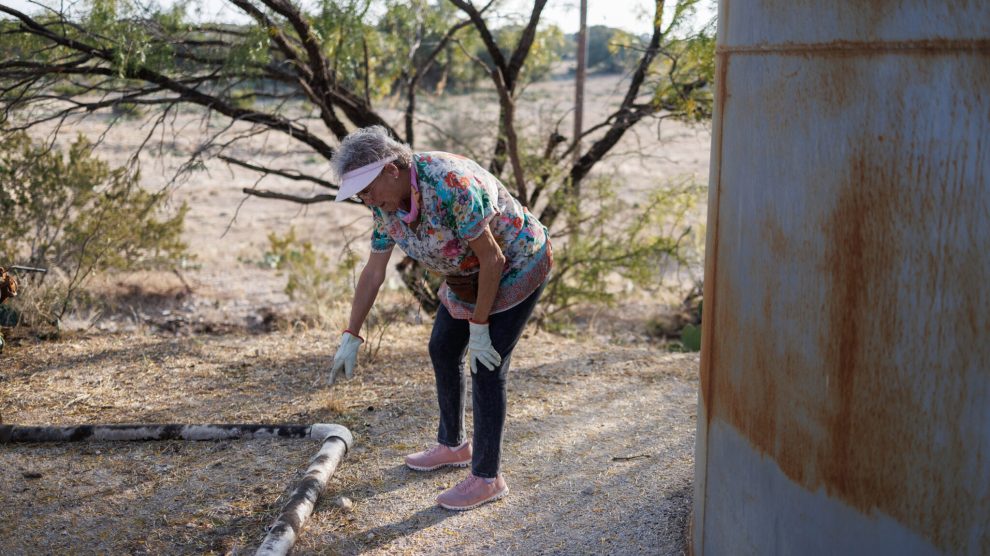 A woman in a floral shirt points out a rusted pipe on the ground next to a rusted tank.