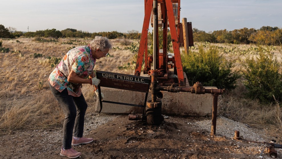 An older woman bends over a piece of oil-pumping machinery on a dry, grassy landscape.