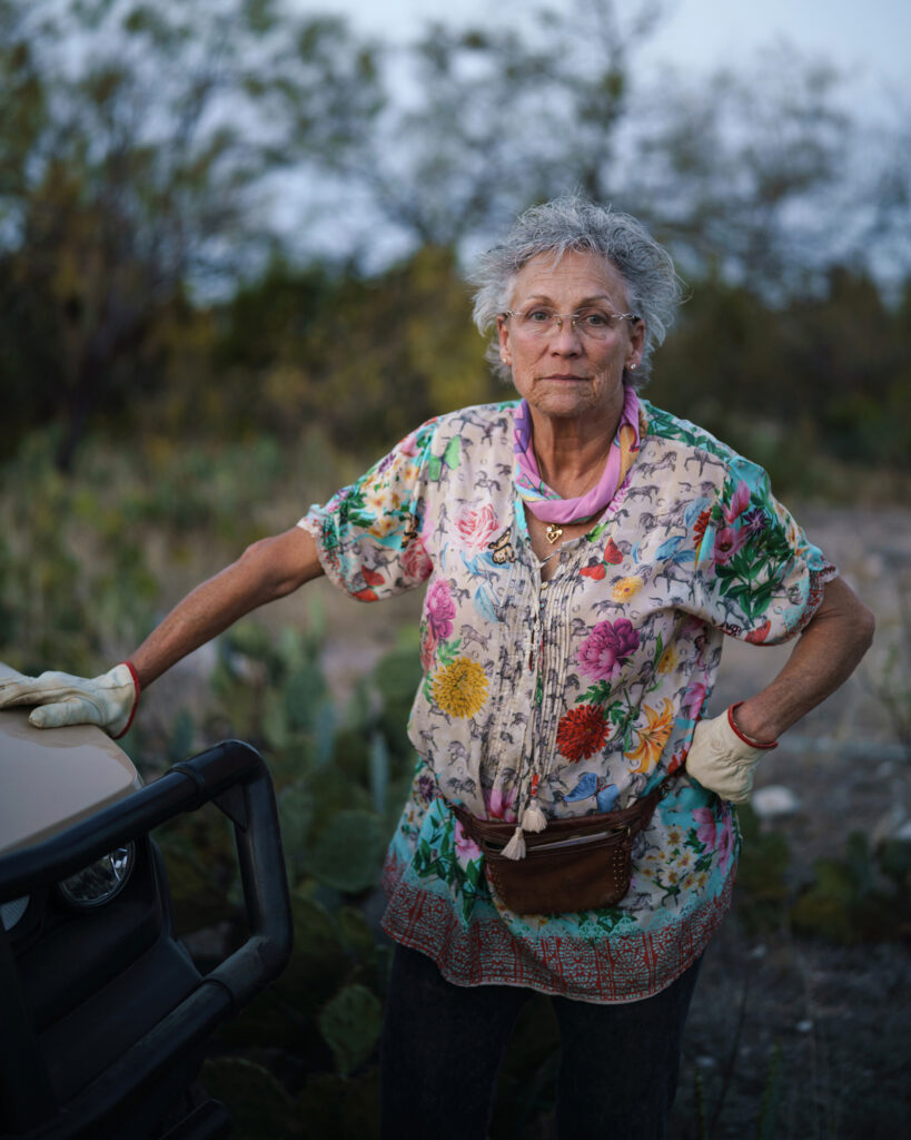 A woman in a floral shirt and gardening gloves with short grey hair looks directly at the camera, standing in a field with her hand on the hood of a truck.