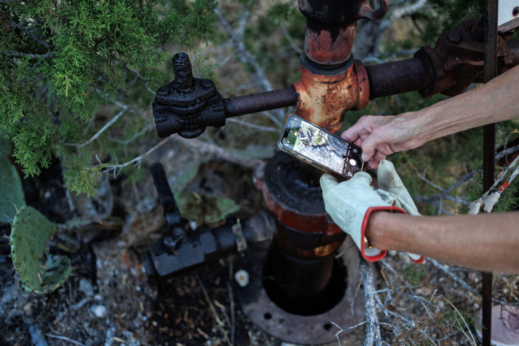 Hands with gardening gloves hold a phone over a well, which is black with oil.