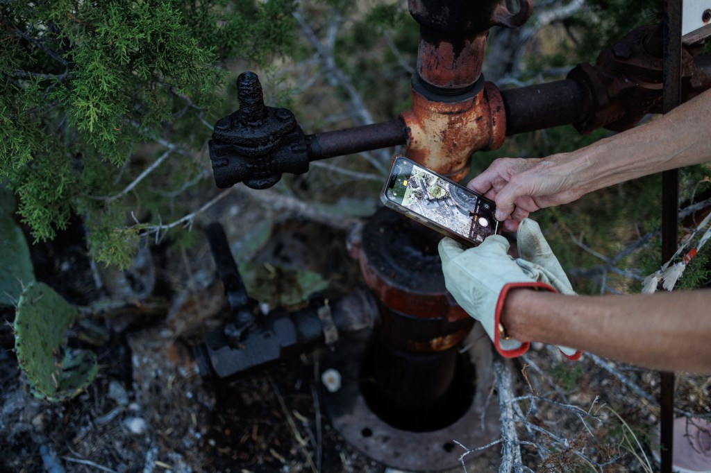 Jackie Lynn Chestnutt photographs a leaky oil well on her property in Knickerbocker, Texas on Tuesday, November 18, 2025. A large number of orphan oil wells are dotted around her property, causing worry that ground water could be contaminated by leeching oil, produced water or chemicals.