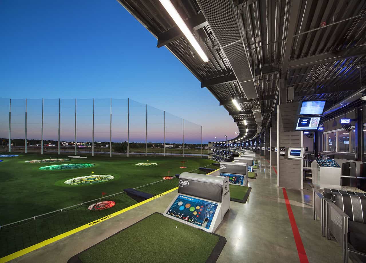 Golf driving range with electronic hitting bays and city skyline at sunset.