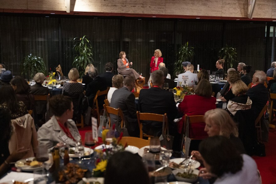 Two women sit on stools speaking into a microphone as a crowd, seated at tables, watches. 