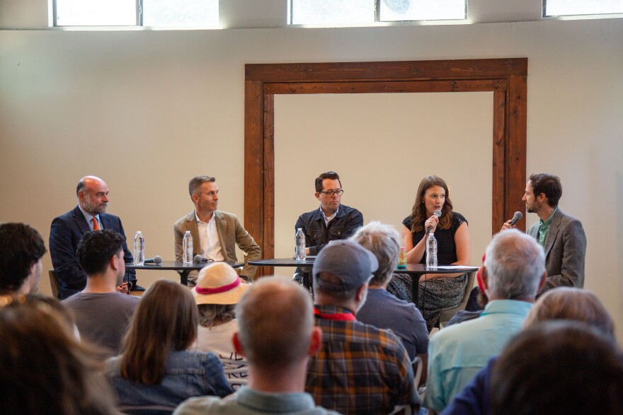 Five people sit at tables in front of a seated audience while two of the panelists speak into handheld microphones.