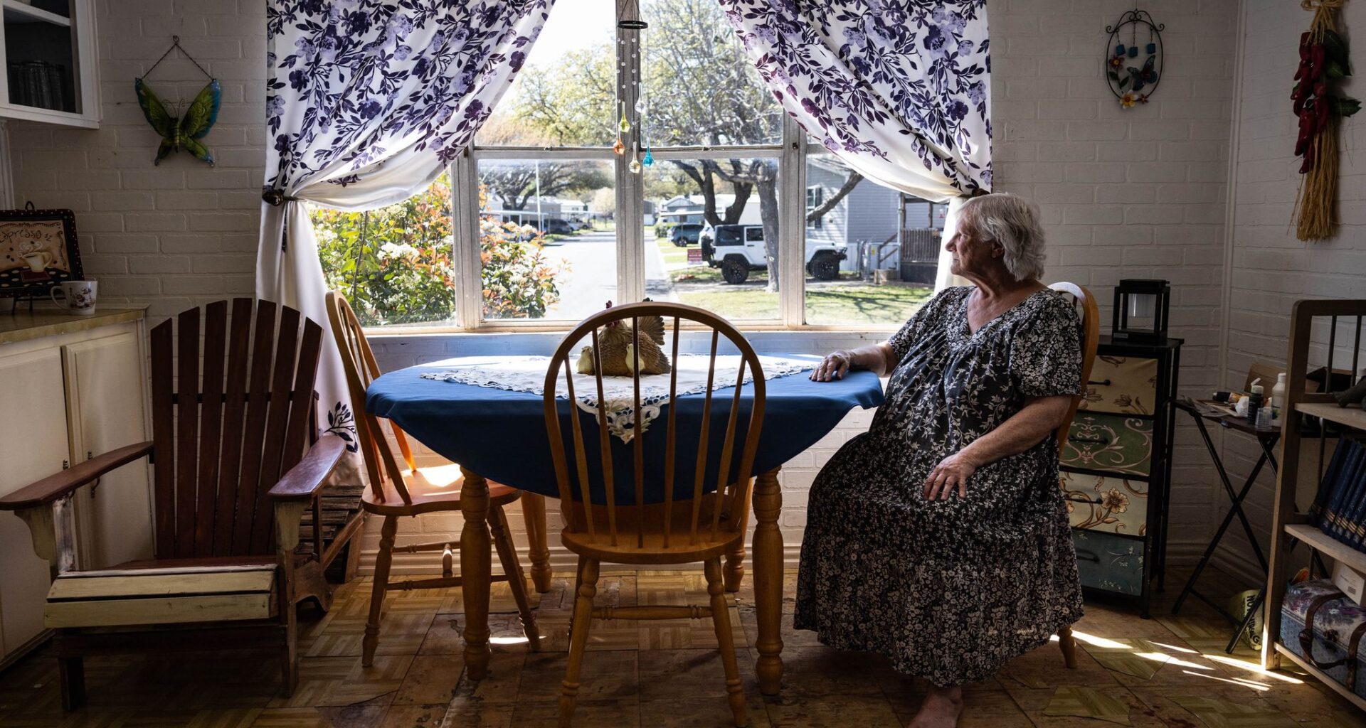 Diane Shields, 77, sits at her kitchen table and looks out the window in Fort Worth on March 20, 2026. Shields said she bought her trailer home 24 years ago because she “fell in love with the windows in the kitchen.” Floor tiles are now peeling away in the kitchen. Years ago, she tried to replace the tiles while making minor repairs to her home. However, they would not come up she said.