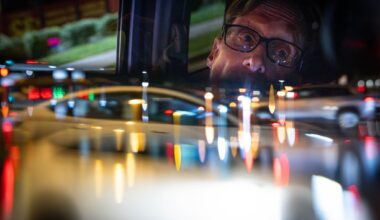 Scott Wheeler is reflected in his rearview mirror while he waits at a stoplight while delivering food in the evening in Dallas, Saturday, April 11, 2026. Traffic in the intersection is obscured using a camera lens filter.