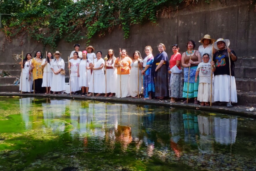 A group of people stand by a spring. Many of them are wearing white. Some of them are holding staffs.