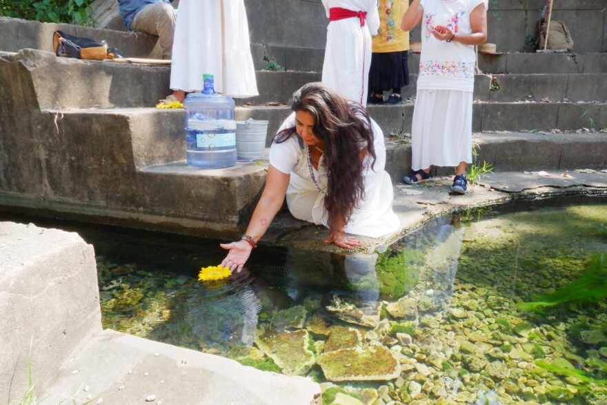 A woman dressed in white kneels next to a spring. Her hand is open next to a yellow flower in the water.
