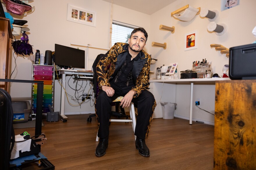 A person dressed in drag king make up poses for a photo in their makeup room.