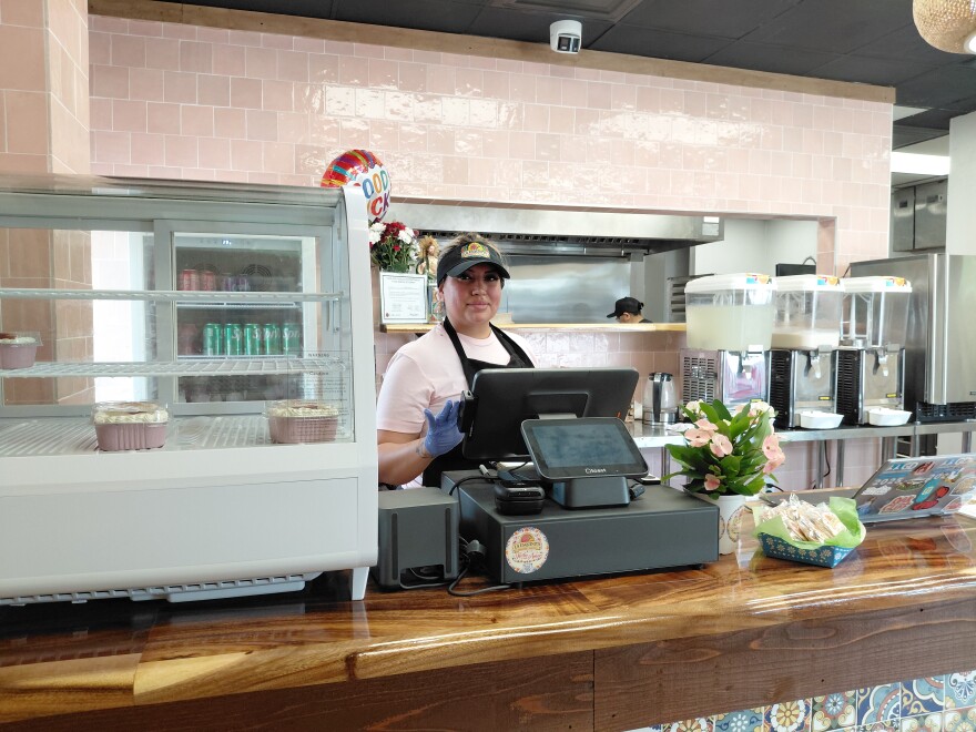 A woman wearing a pink shirt, black apron and black baseball cap stands behind the counter of a bakery,
