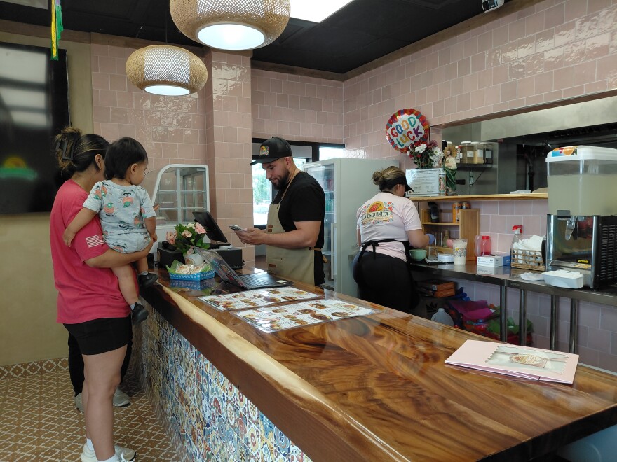 A woman carrying a baby is at a restaurant counter. Behind the counter are a man and woman working in the restaurant.