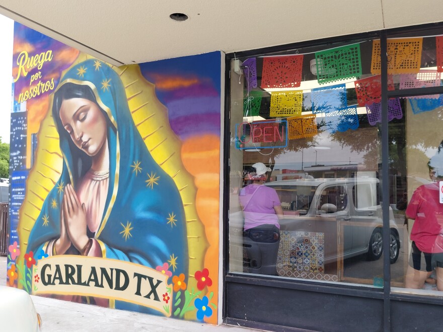 A colorful mural on the side of a building shows the Virgin of Guadalupe with the words "Ruega por nosotros" above her and "Garland TX" below. Next to her the inside of a bakery with customers waiting around is visible through a window.