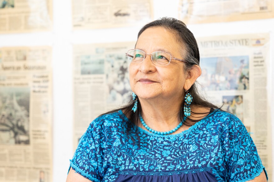 A woman wearing aqua green jewerly and a top is shown sitting in front of newspaper front pages.