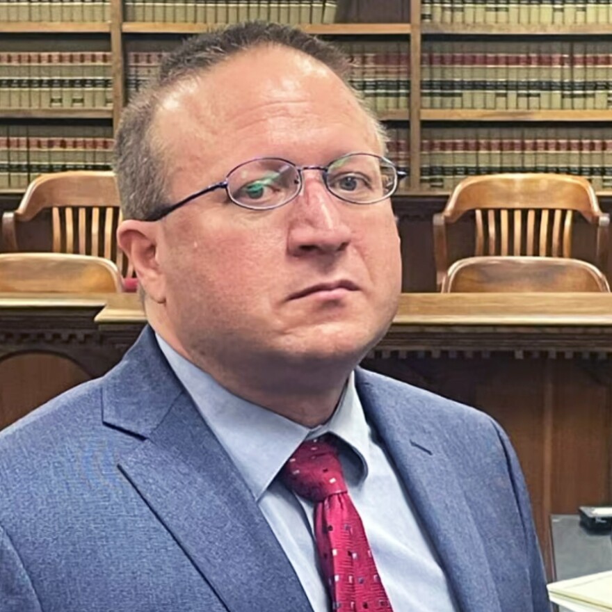 A man wearing a suit and glasses sits in a courtroom.