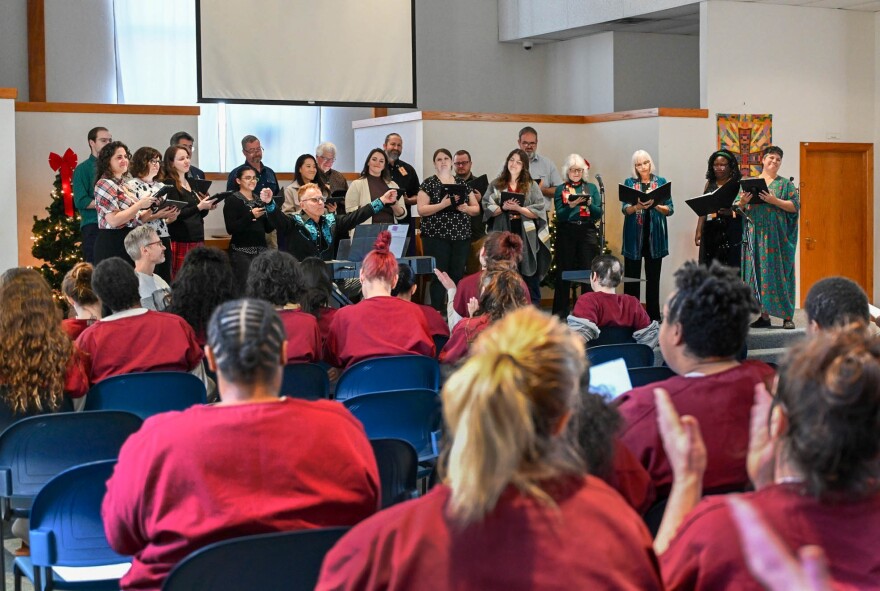 Female inmates seated in the audience clap and look towards the Conspirare chorus singers and conductor standing near the, at the front of a chapel.