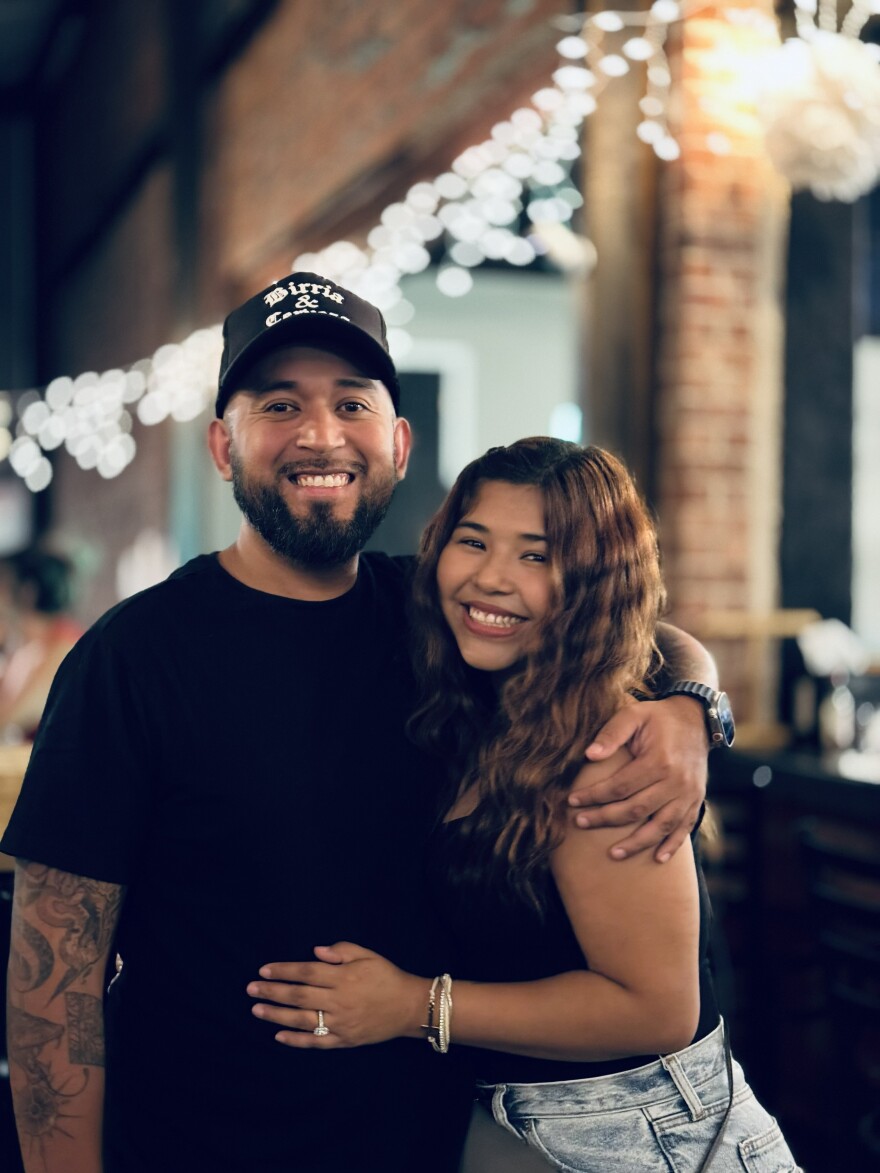 A man in a black shirt and ballcap poses with his arm around a woman in a black top and jeans. They are standing in a street with lights behind them and both are smiling. 