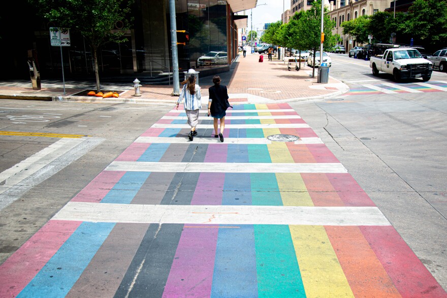 Pedestrians walking across a rainbow crosswalk. The crosswalk includes transverse white lines.