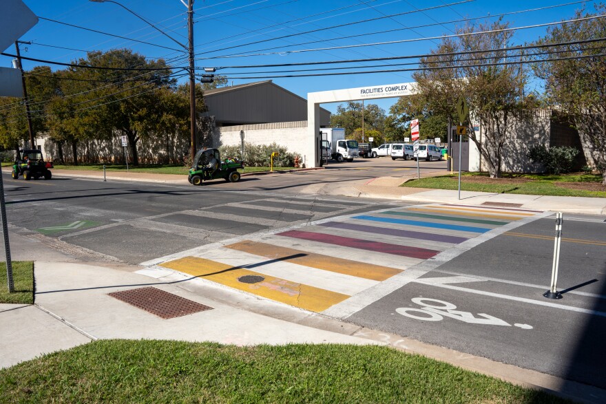 A rainbow crosswalk at Manor Road at Leona Street in Austin, Texas on Tuesday, Nov. 11, 2025. A sign reading "Facilities Complex" is visible as green golf cart-type vehicles drive out.