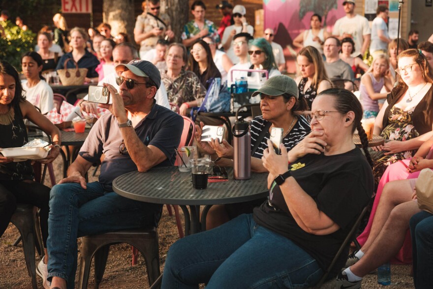 The audience sits at tables, watching and recording performances during the Here Be Monsters festival in 2025.