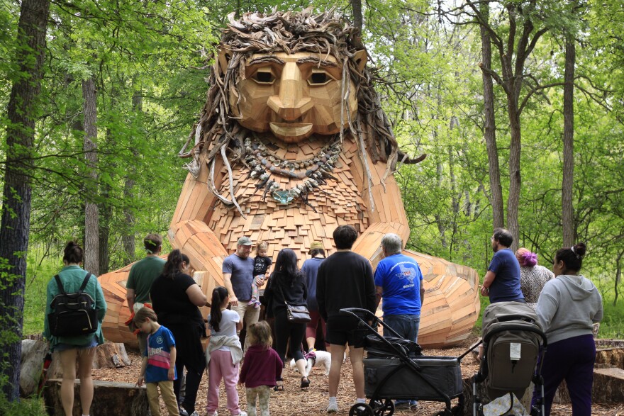 A giant wooden troll sculpture in the woods. Around a dozen people are standing in front of the troll, observing it.