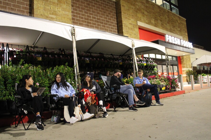 Customers chat with one another while waiting. Behind the row visible here, another group of people are waiting under the awning. May 19, 2026.