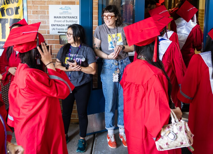 Teenagers dressed in red caps and gowns walk into a building. Two women stand on the back looking excited