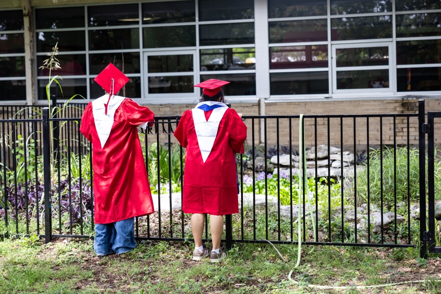 The back of two senior students wearing a red cap and gown look over a fence
