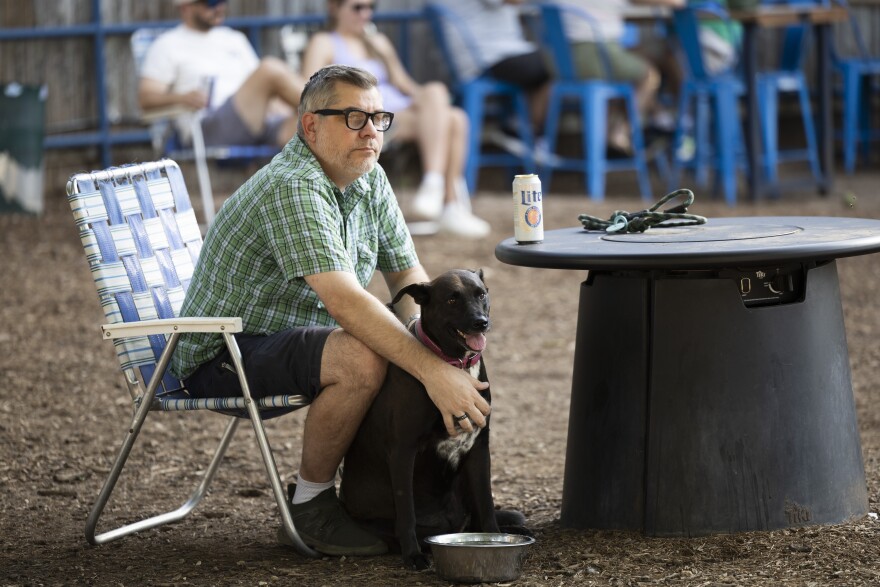 A man pets a dog as he sits in a lawn chair at a small table at an outdoor bar.