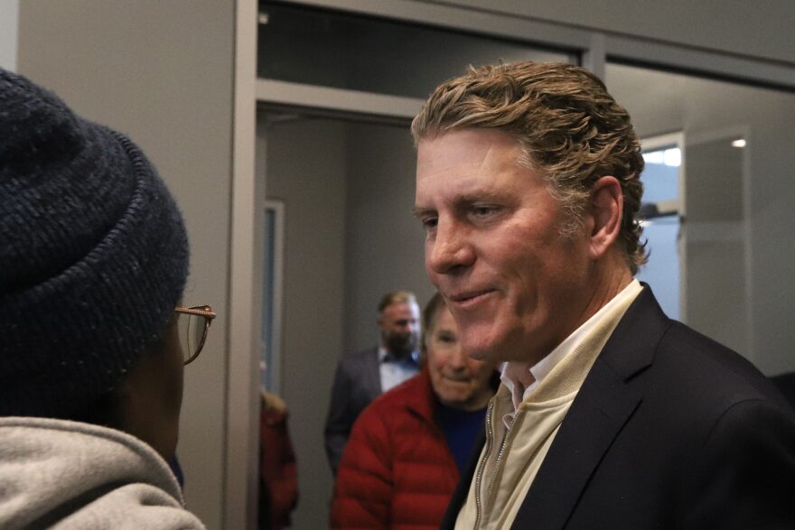 A smiling man in a suit speaks to a person in glasses and a knit hat.