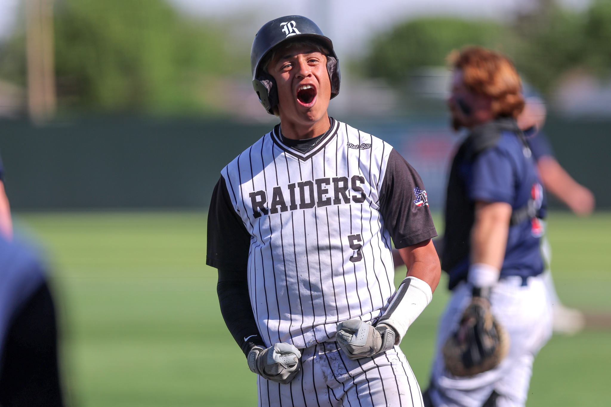 Baseball player in pinstripe Raiders jersey shouting with mouth open, wearing helmet on a sunny field, holding a bat.