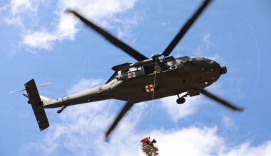 A person and a dog sit in a suspended basket lift attached to a helicopter that is airborne during a search and rescue exercise.