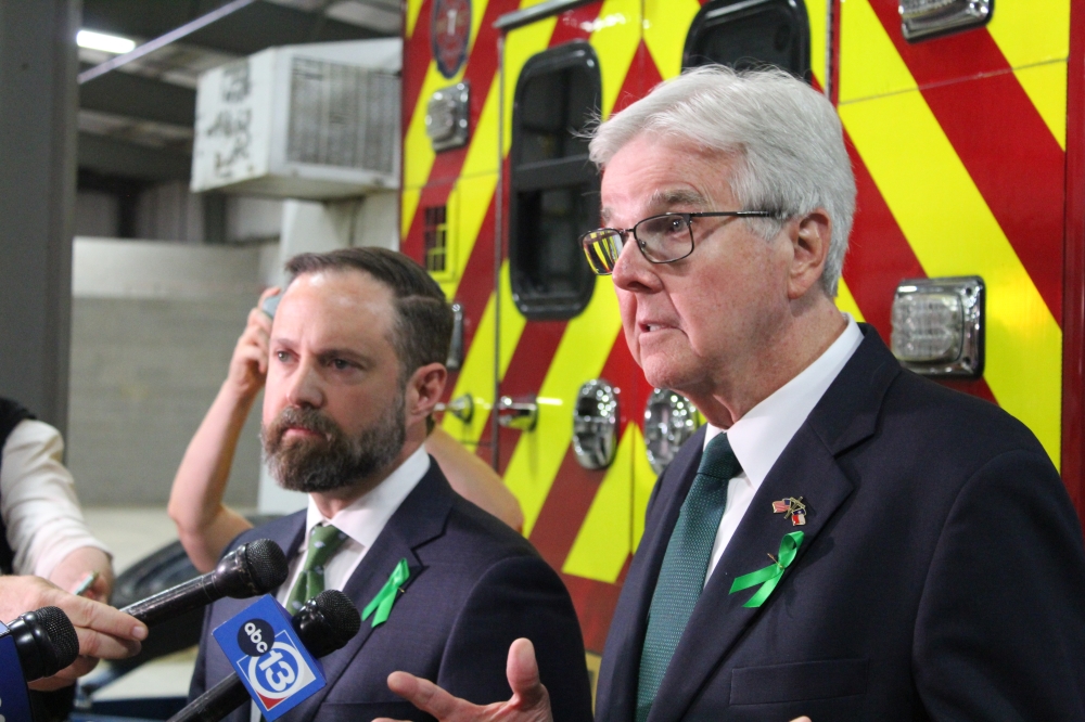 Dustin Burrows and Dan Patrick speak to reporters in front of a red and yellow striped emergency vehicle in Kerrville.