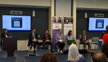 Five professionally dressed people, including three men, two women and one empty chair, sit at the front of a room during a panel, with another woman standing at a podium and one ore at a microphone positioned at the front of the room. The backs of four audience member's heads are in frame. A QR code sits on two screens behind the panel members, with text text that reads