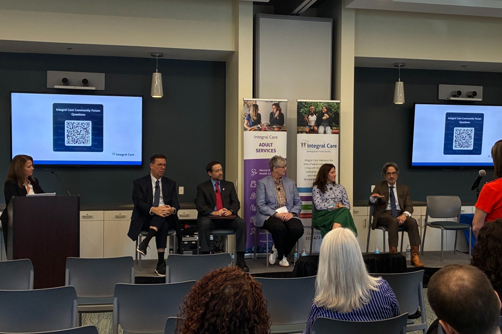 Five professionally dressed people, including three men, two women and one empty chair, sit at the front of a room during a panel, with another woman standing at a podium and one ore at a microphone positioned at the front of the room. The backs of four audience member's heads are in frame. A QR code sits on two screens behind the panel members, with text text that reads