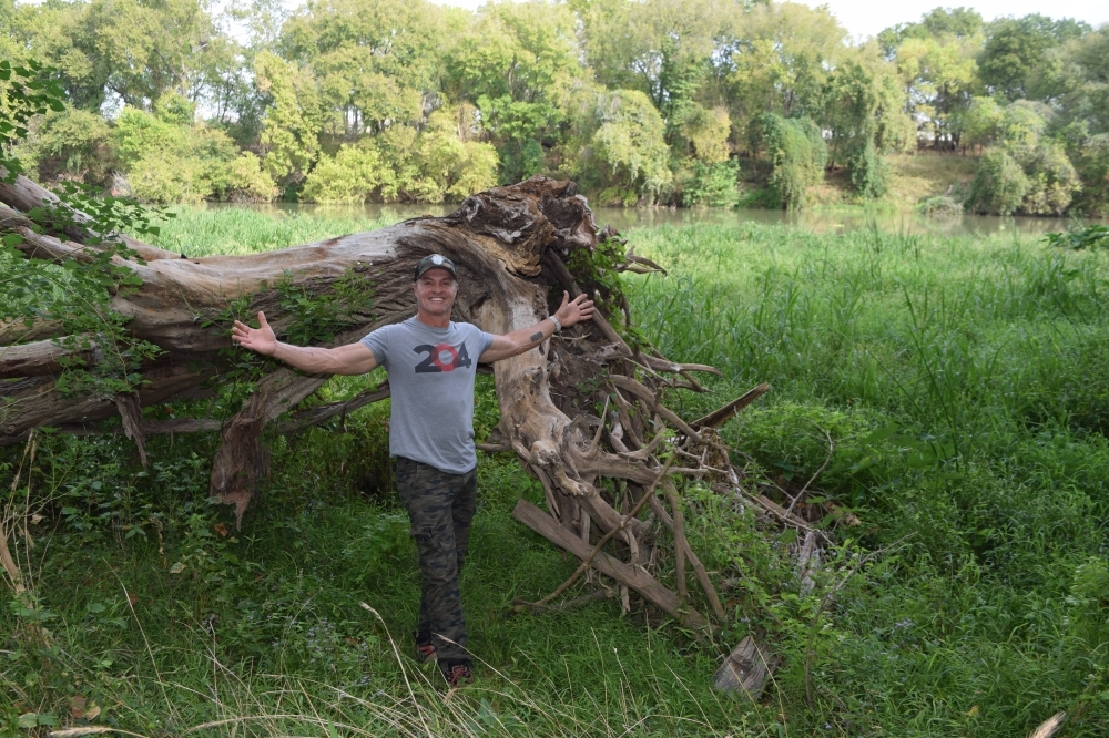 The owner stands in front of a large uprooted tree on the property surrounded by green trees and grass.