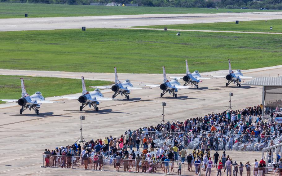 A crowd of people watch as a line of aircraft move down a tarmac.
