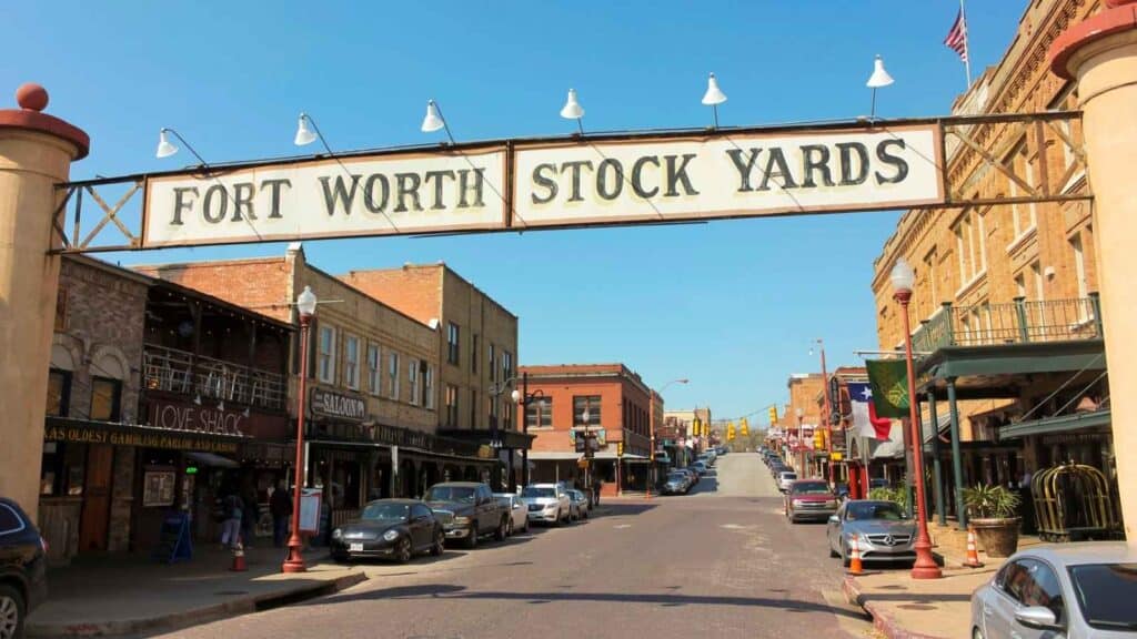 A street view of the Fort Worth Stock Yards, showing historic buildings, parked cars, and a large overhead entrance sign.