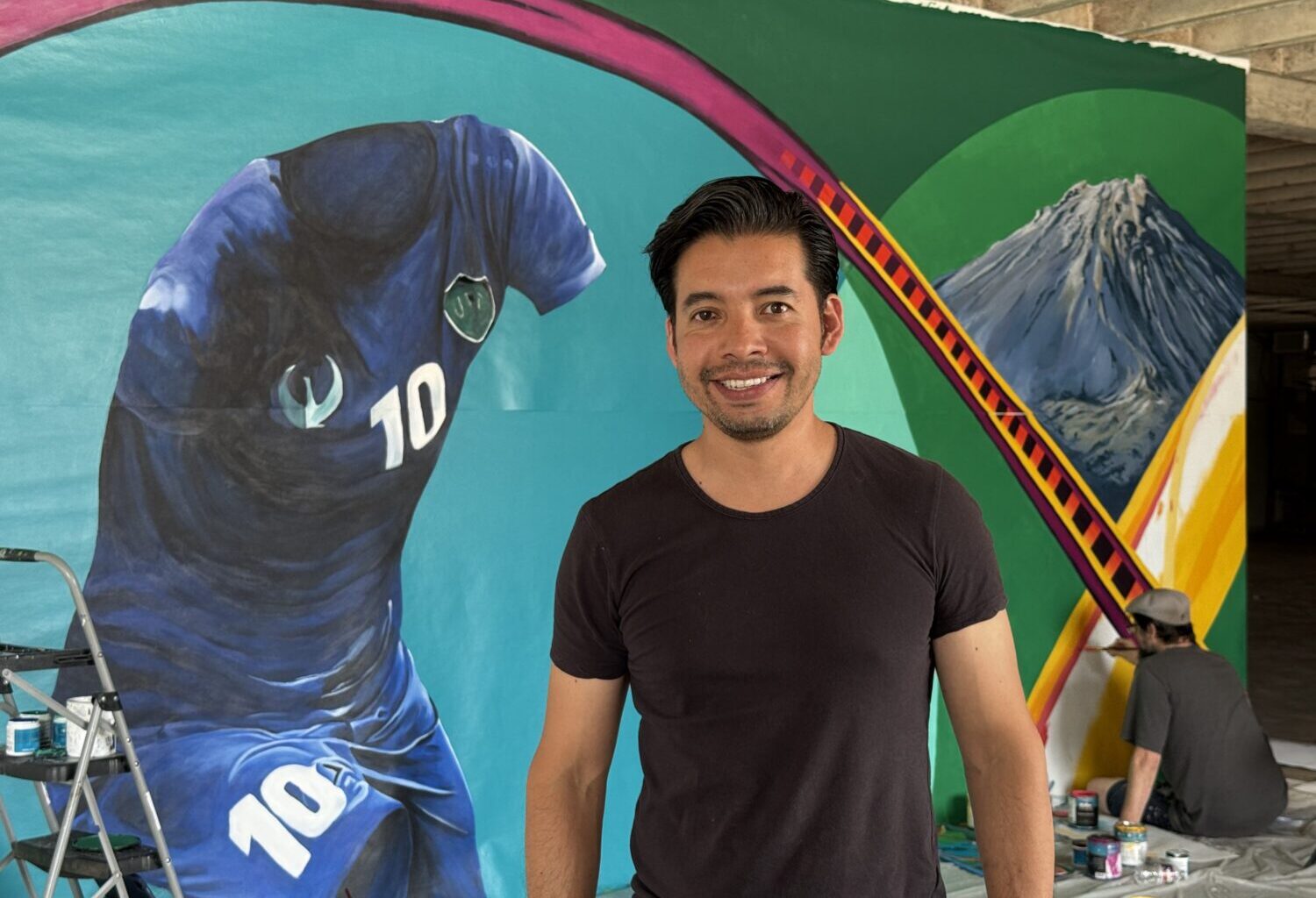 Betirri smiles in front of the Gateway Mural in his studio