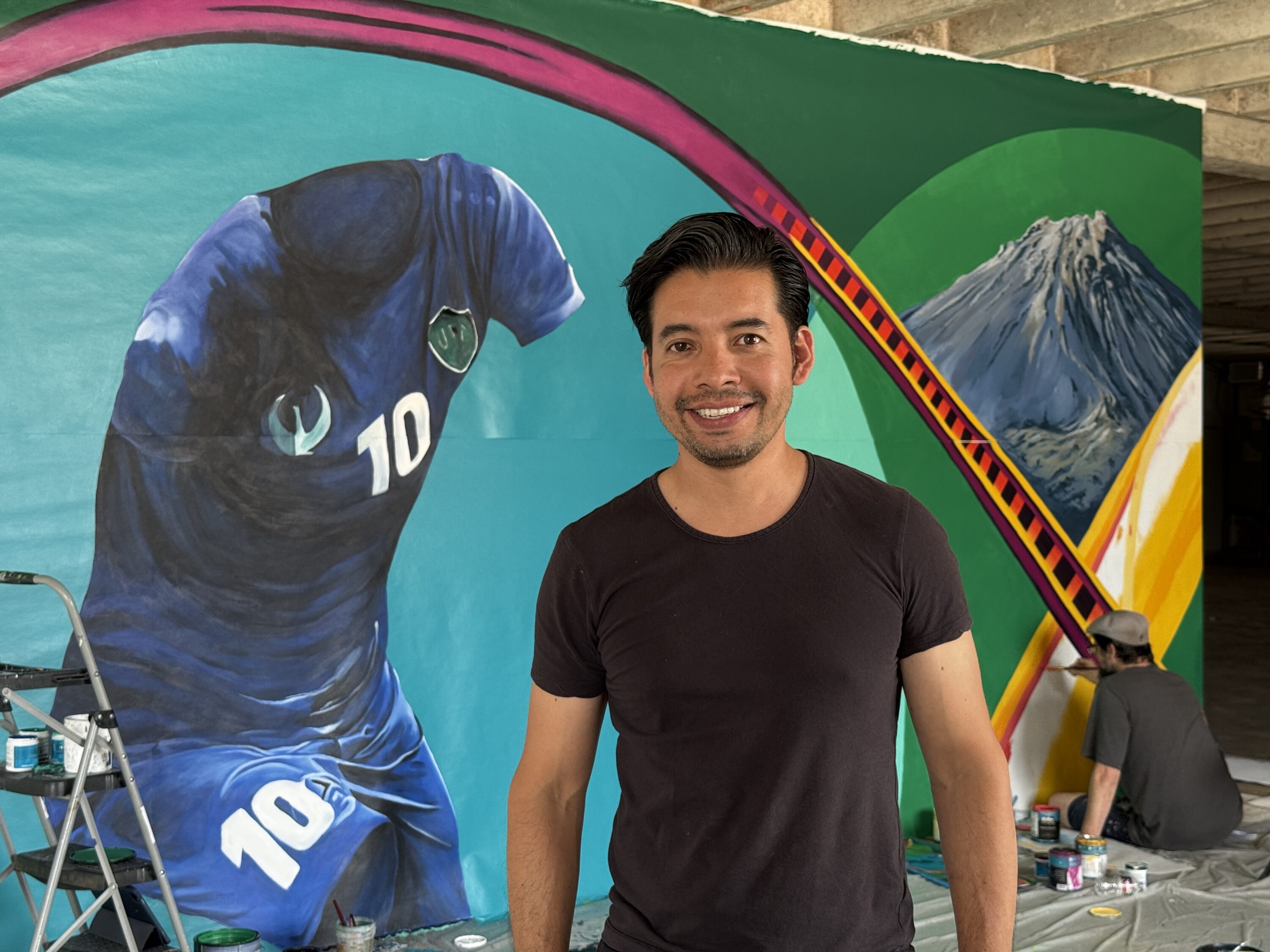 Betirri smiles in front of the Gateway Mural in his studio