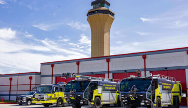 Three airport fire and rescue vehicles parked in a row outside a fire station building, with a tall air traffic control tower rising behind them. The vehicles are painted in bright yellow and black with ‘Fire & Rescue’ markings, and the station has large red bay doors under a partly cloudy sky.