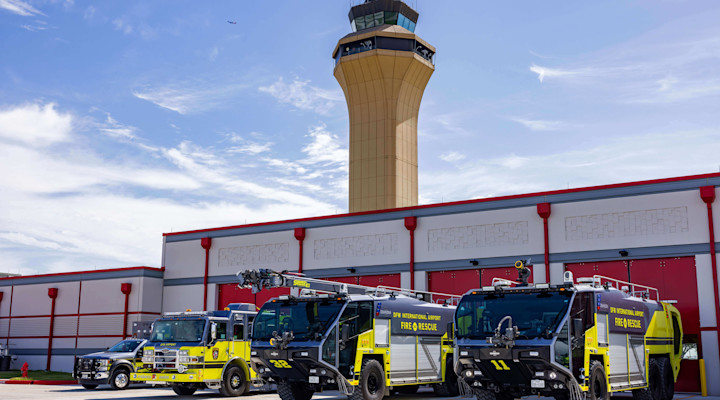 Three airport fire and rescue vehicles parked in a row outside a fire station building, with a tall air traffic control tower rising behind them. The vehicles are painted in bright yellow and black with ‘Fire & Rescue’ markings, and the station has large red bay doors under a partly cloudy sky.