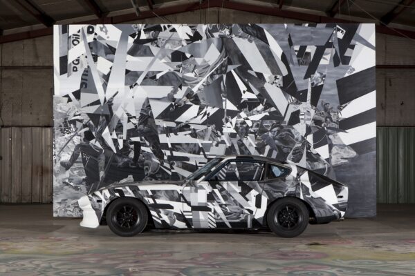 A sportscar in front of a large wall painting, both painted identically in fragmented black and white patterning, in a large exhibition space.