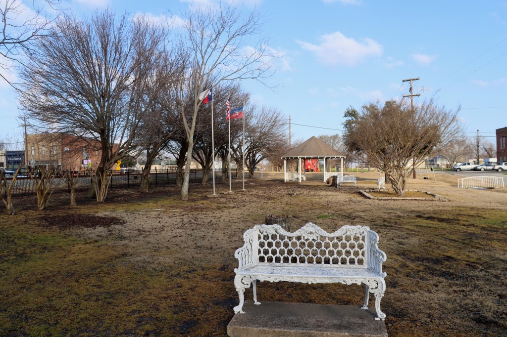 A white ornate metal bench in the foreground of a park with bare trees, a white gazebo, and flags under a partly cloudy sky.